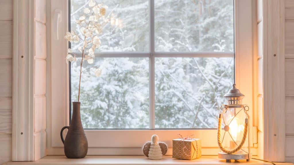 Fönster med vit träram, snötäckta träd utanför och en lampa som lyser i fönsterkarmen